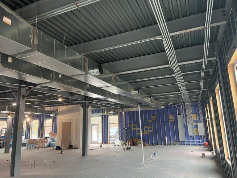 A large industrial room under construction with exposed ductwork and wiring on the ceiling, partially finished walls, and construction materials scattered on the floor.
