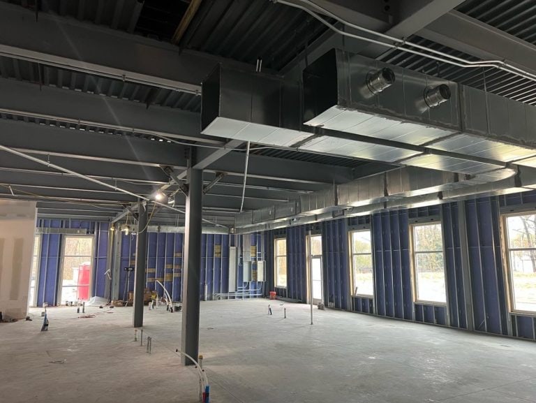 A large, partially constructed room with exposed ductwork and electrical wiring. Metal framing and unfinished walls are visible, with some construction materials on the floor.