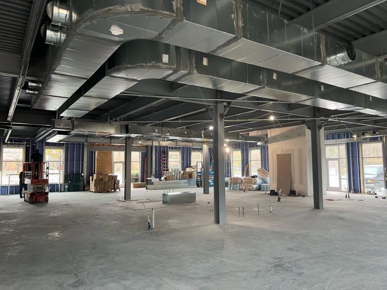 Large, open industrial space under construction with exposed ductwork on the ceiling, concrete floor, and materials stacked in the background.