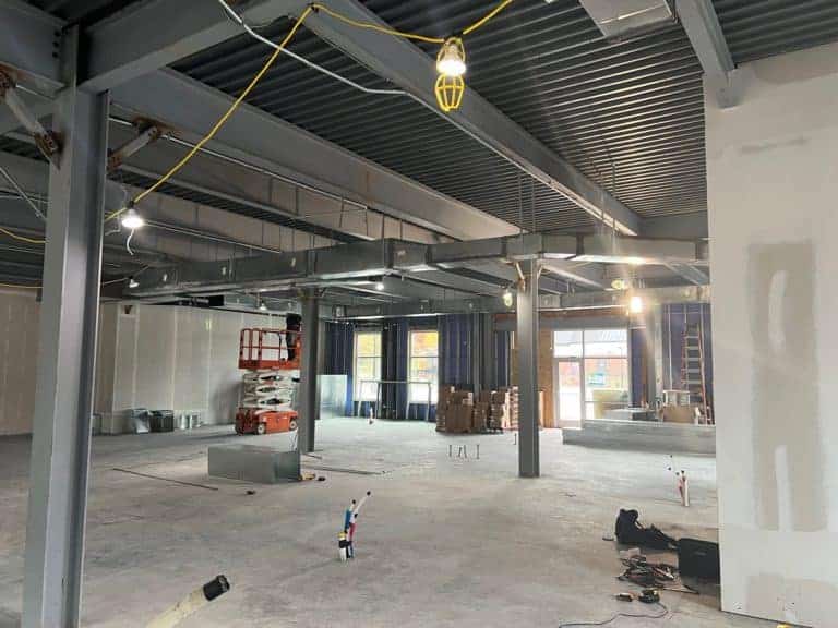 A large, unfinished interior space with exposed steel beams, electrical wiring, and construction materials scattered. A scissor lift and boxes are visible in the background.