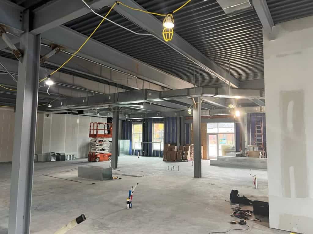 A partially constructed interior of a commercial space with exposed metal beams, wiring, construction materials, and machinery.