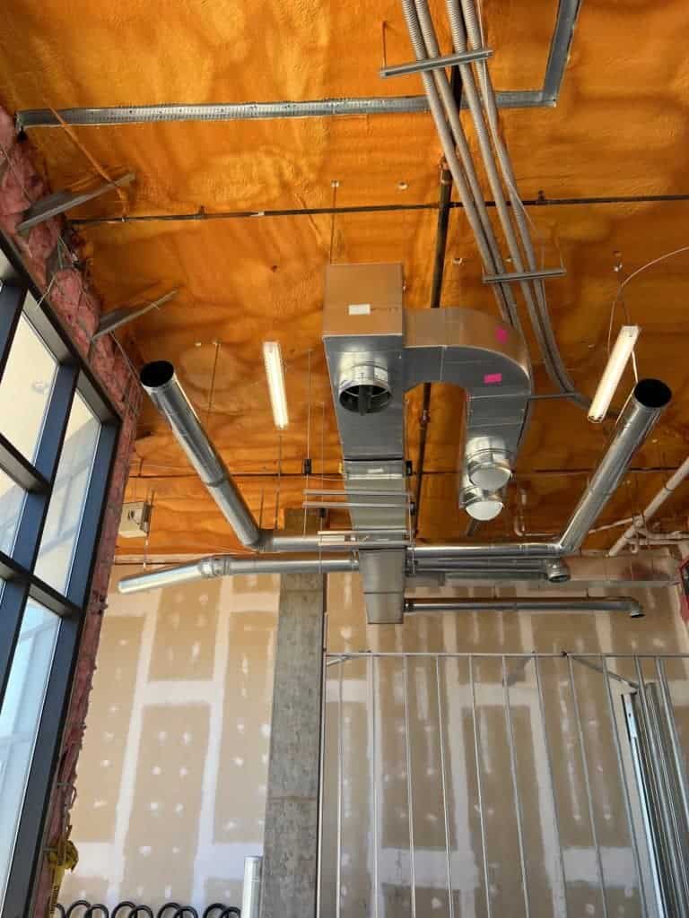 Exposed HVAC ducts and electrical conduits run along the ceiling of a construction site with partially finished walls and insulation.