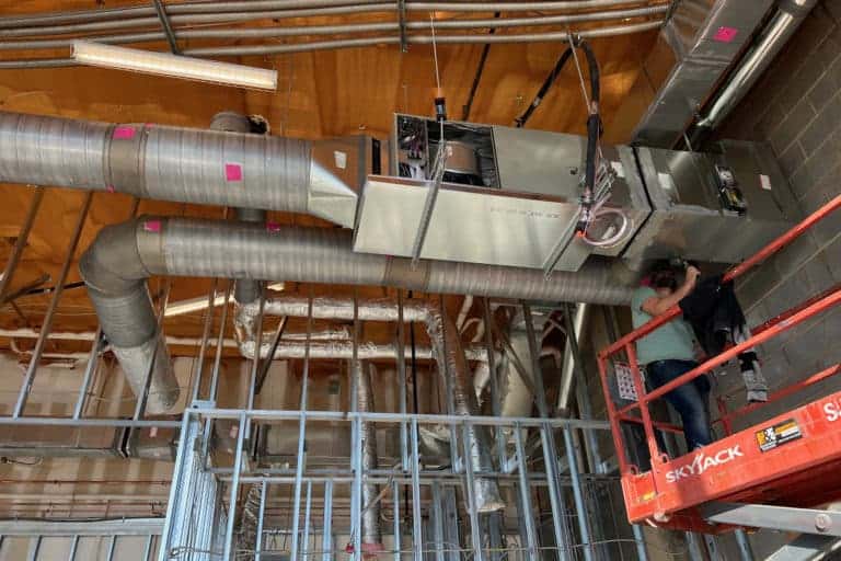 A worker on a lift installs or inspects HVAC ductwork and equipment in a construction area with exposed wiring and framework.