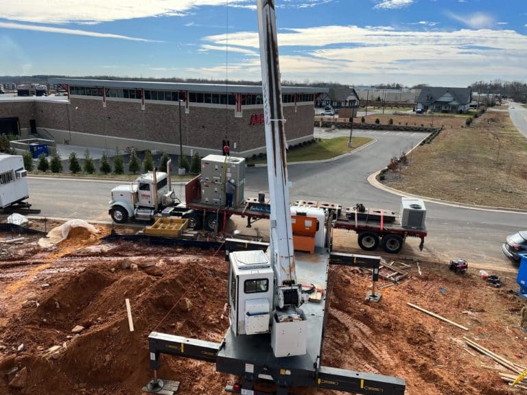 Construction site with a large crane positioned in a dug-out area. Trucks and equipment are nearby on a paved road. In the background, there is a commercial building and residential homes.