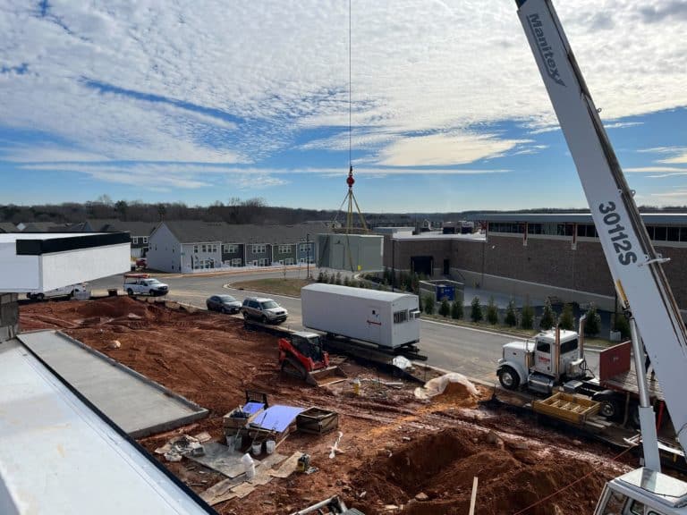 Construction site with cranes lifting materials, trucks, and construction equipment visible. Nearby, there are buildings, and the sky is partly cloudy.