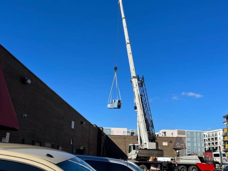 A crane hoisting a large piece of equipment above a parking lot, with a clear blue sky and buildings in the background.
