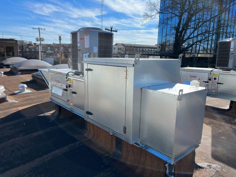 A large industrial HVAC unit sits on the flat roof of an urban commercial building, with other similar units and a clear sky in the background.