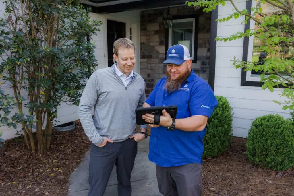 Two men stand outside a house, looking at a tablet held by the man in a blue shirt and cap. The man in a gray sweater smiles as he observes the tablet. Shrubs and trees are visible around them.