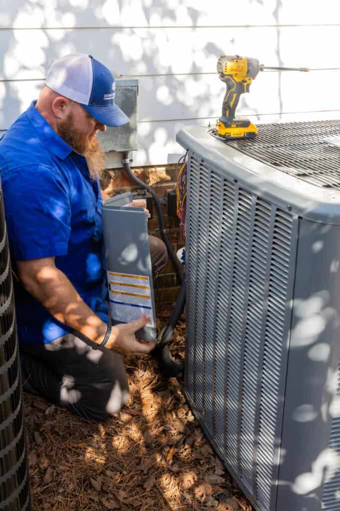 Technician in a blue shirt and cap works on an outdoor air conditioning unit, holding a part while a yellow drill rests on top of the unit.