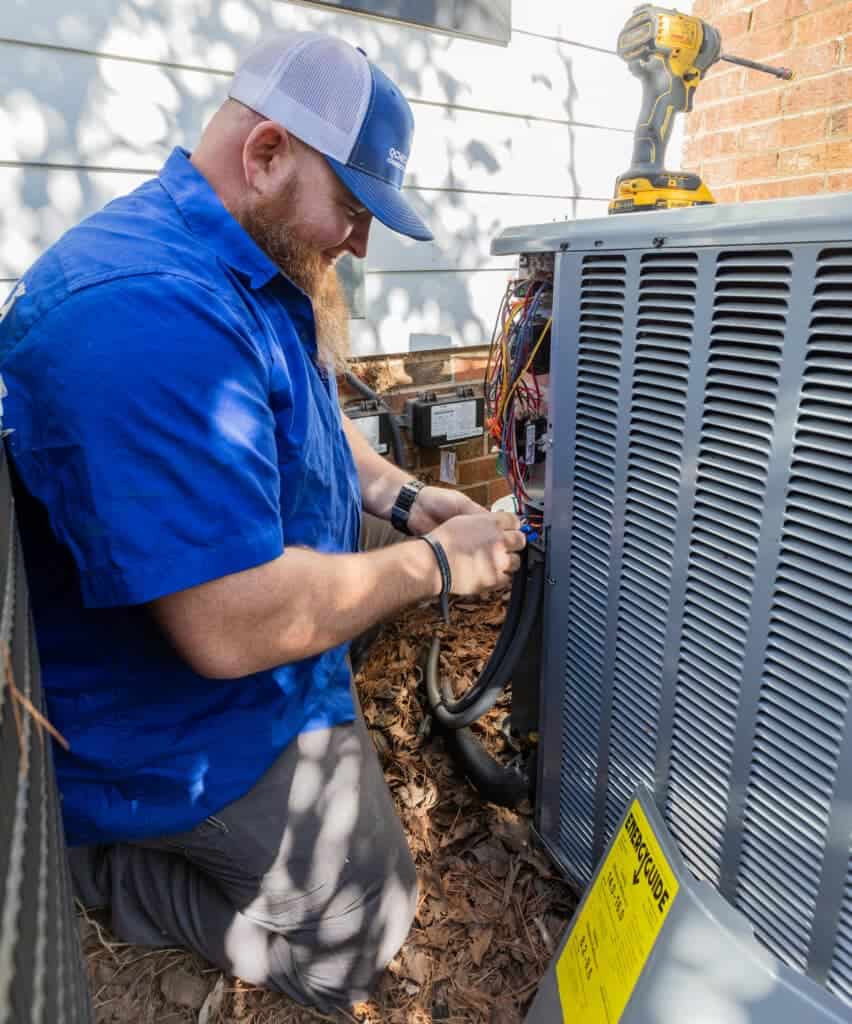 A technician in a blue shirt works on an air conditioning unit outdoors. A yellow drill is placed on top of the unit.