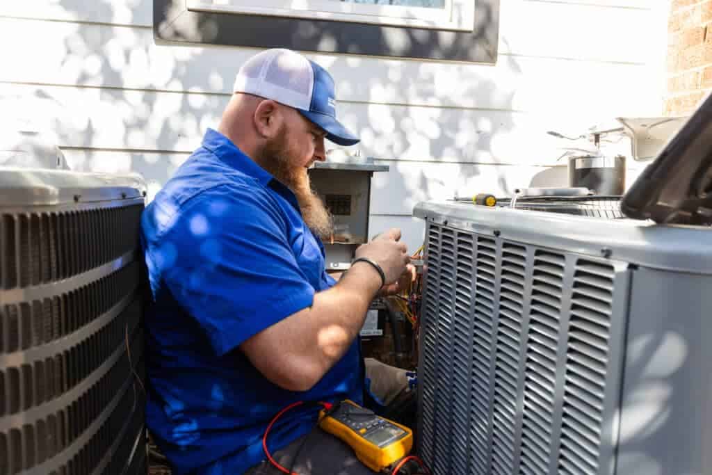 A technician with a long beard wearing a blue shirt and cap is repairing an air conditioning unit outdoors between two large AC units, with his tools and a digital multimeter nearby.