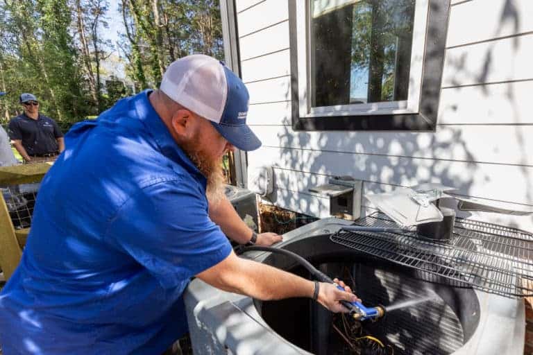 A man in a blue shirt and cap uses a water hose to clean an outdoor air conditioning unit. Another person stands watching in the background.