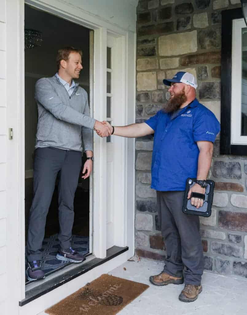 A man in a gray sweater stands in the doorway of a house shaking hands with another man in a blue shirt and cap holding a tablet.