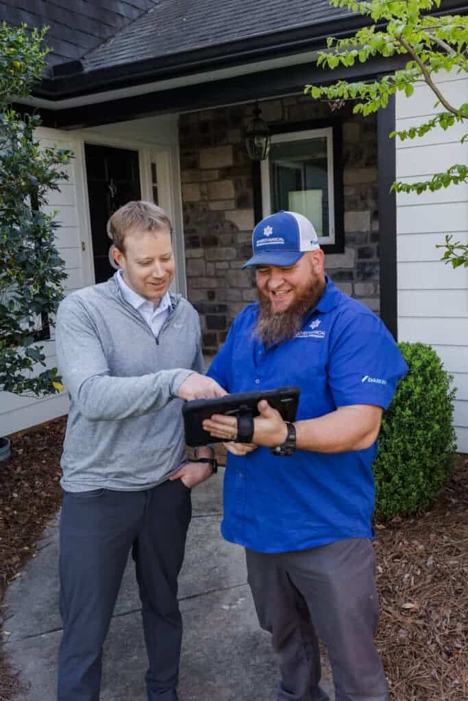 Two men standing outside a house, one wearing a blue shirt and cap holding a tablet, and the other in a gray sweater and dress shirt looking at the tablet.