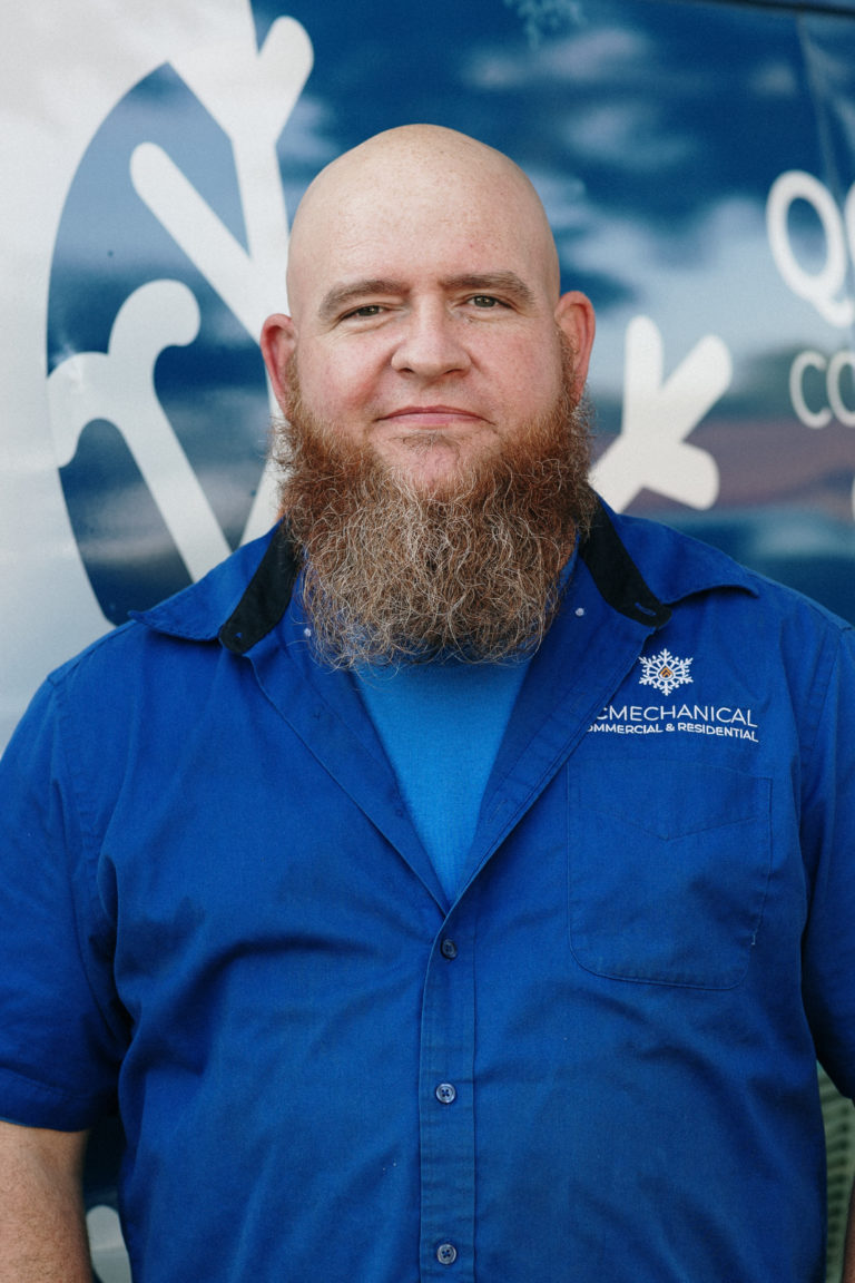 A bearded man in a blue shirt stands in front of a vehicle with a logo.