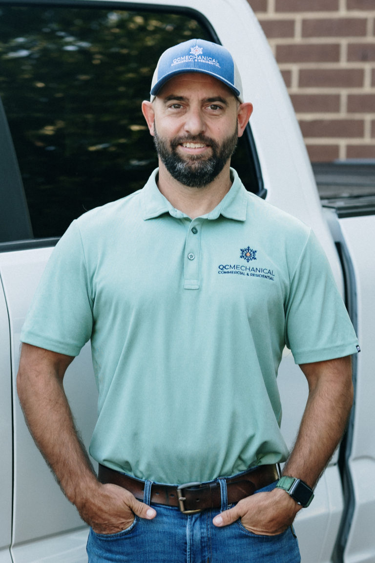 Man with a beard wearing a green polo and a cap stands in front of a white vehicle and brick wall.