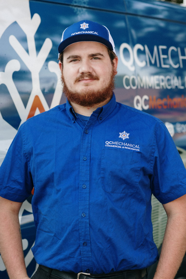 A person wearing a blue QC Mechanical uniform and cap stands in front of a company van with QC Mechanical branding.