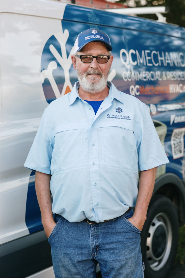 Man in QC Mechanical uniform stands in front of a company van with hands in pockets, wearing a cap and glasses.