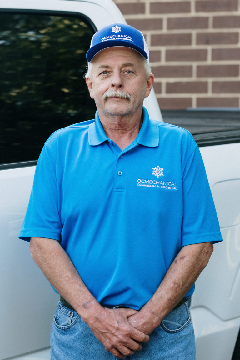 Man in a blue OC Mechanical polo shirt and cap stands in front of a white vehicle, with a brick wall in the background.