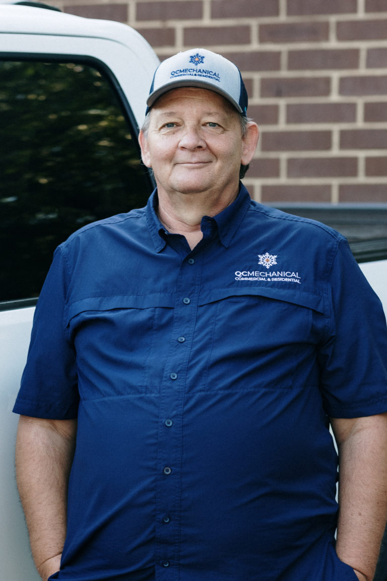 Man in a blue shirt and cap stands by a white vehicle, with a brick wall in the background.