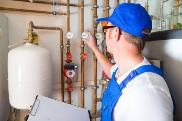 A worker in a blue uniform checks gauges on pipes in a mechanical room, holding a clipboard to ensure how the heating system works.