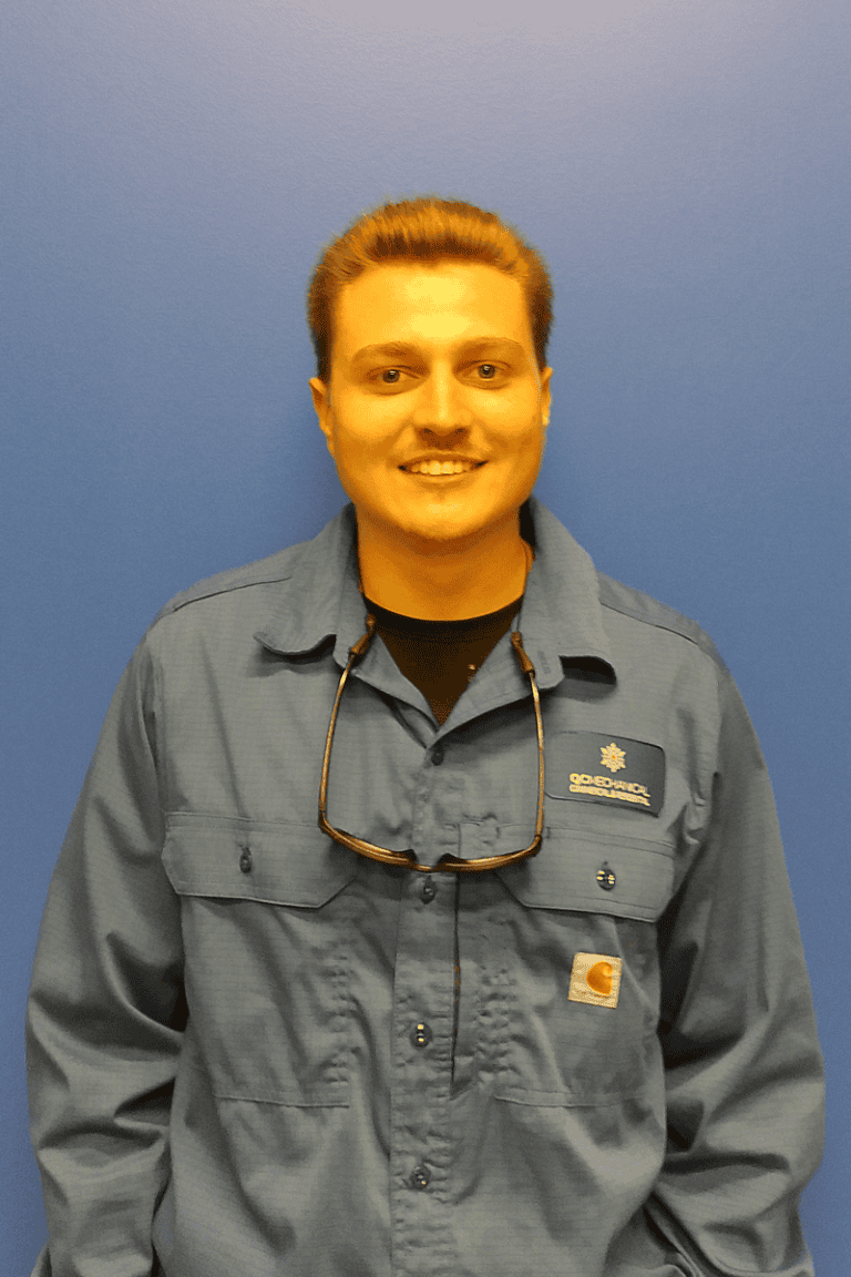 A person with short hair smiles at the camera, wearing a blue collared work shirt with a name tag and sunglasses hanging from the collar, standing against a blue wall.