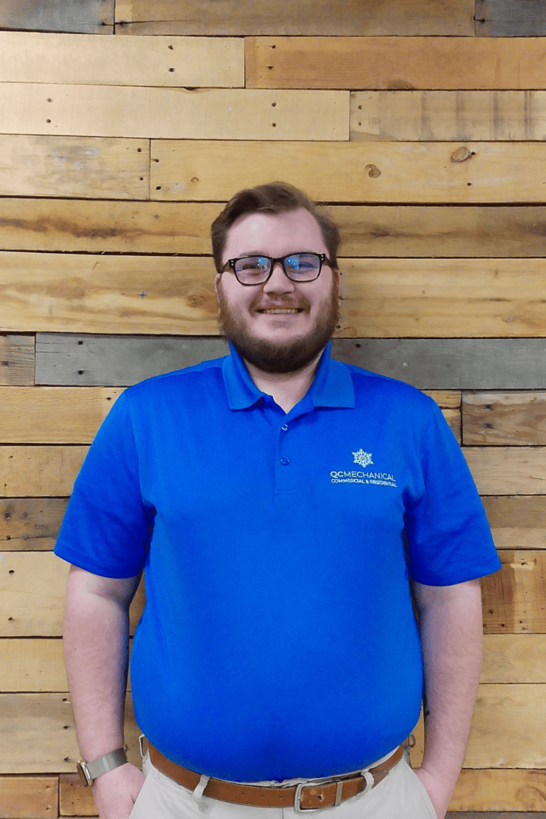 A man with glasses and a beard, wearing a blue polo shirt and khaki pants, stands in front of a wood plank wall, smiling at the camera.