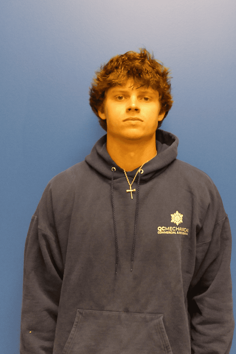 A young man with brown hair wearing a dark blue hooded sweatshirt and a cross necklace stands against a plain blue wall.