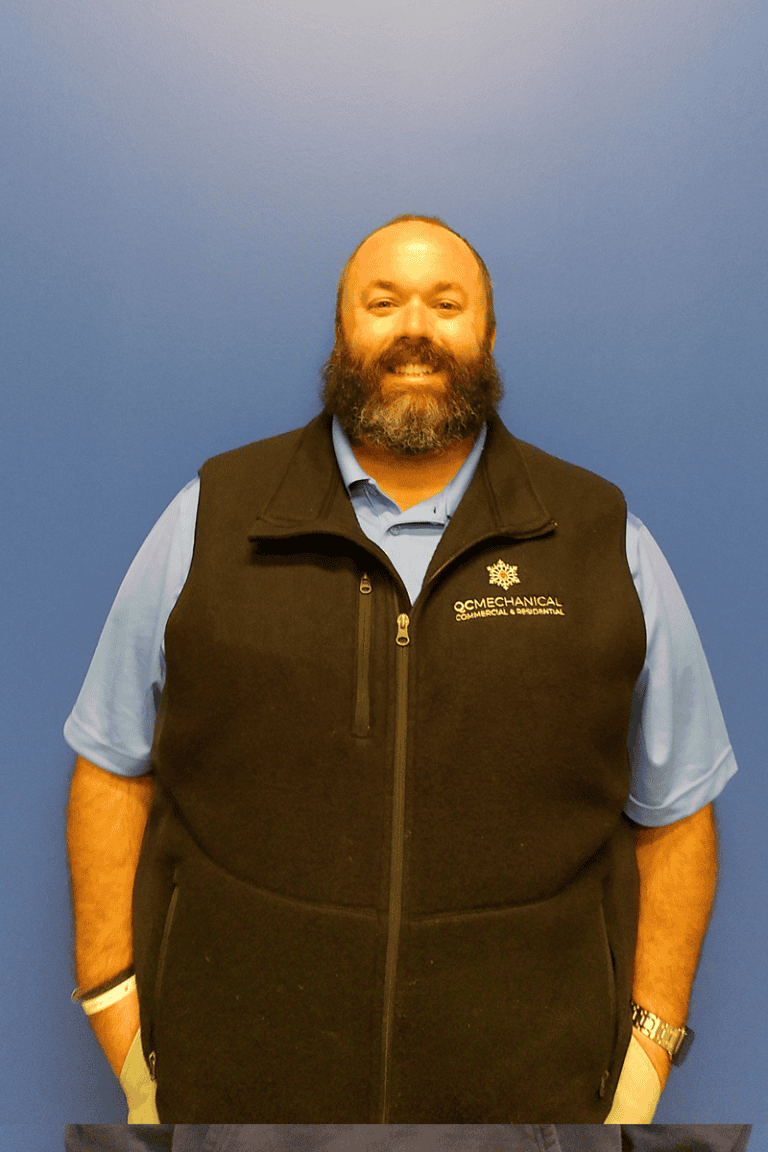 A man with a beard stands against a plain blue wall, wearing a light blue shirt and a black vest with an