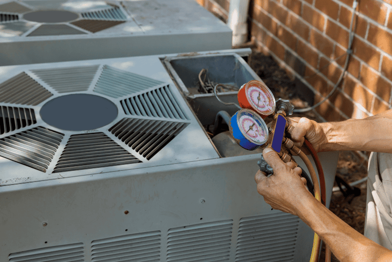 A person checks the pressure on an air conditioning unit using a gauge manifold set outdoors next to a brick wall.