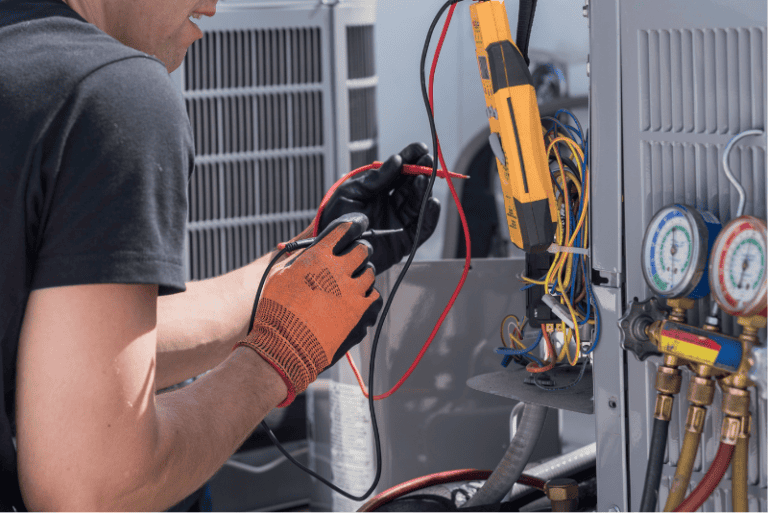Technician wearing gloves uses tools and measures electrical wires inside an open HVAC unit, with gauges and devices visible.