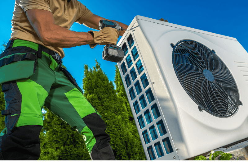 A technician in green and black work pants uses a power drill to install or service an outdoor air conditioning unit on a clear, sunny day.