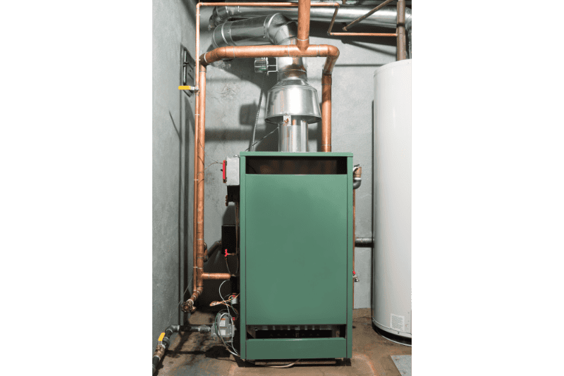 A green residential furnace is installed in a utility room with visible copper piping and a white water heater nearby.