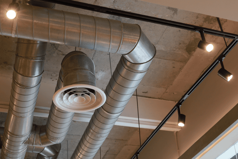 Exposed metal air ducts and a circular vent on a concrete ceiling with track lighting fixtures turned on.