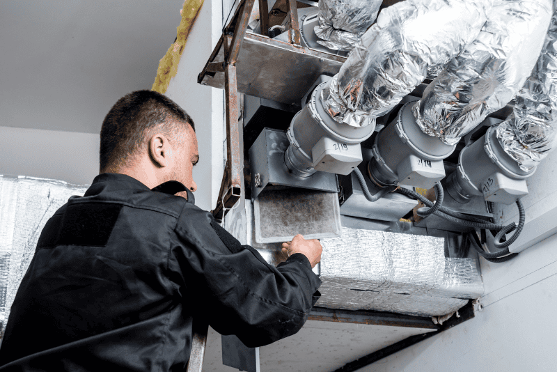A person wearing a black uniform inspects or repairs a large industrial HVAC system with metal ducts and insulated pipes.