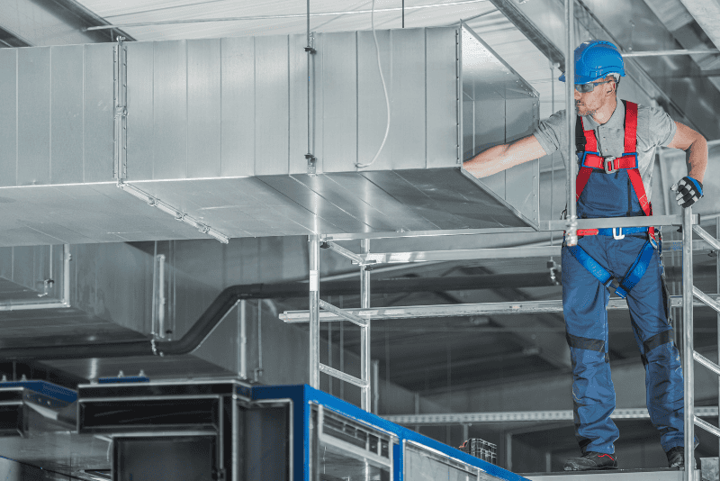 A construction worker wearing safety gear inspects a metal air duct system on a raised platform inside an industrial building.