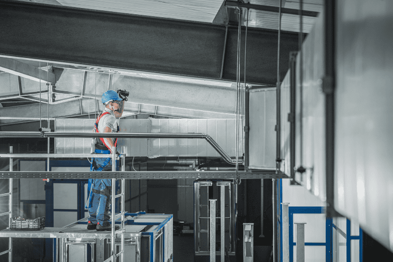 A worker in safety gear and a blue helmet inspects ventilation ducts inside an industrial building.