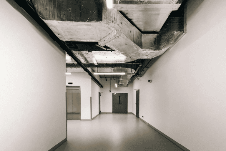 A modern hallway with exposed ventilation ducts on the ceiling, white walls, gray doors, and an elevator on the left.