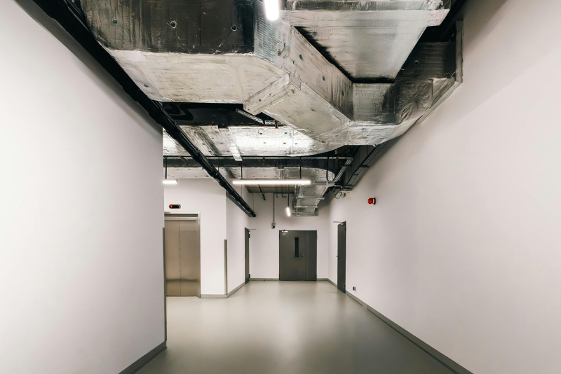 A modern hallway with exposed ventilation ducts on the ceiling, white walls, gray doors, and an elevator on the left.
