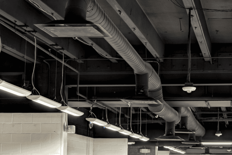 Exposed black ventilation ducts and hanging fluorescent lights on an industrial-style ceiling above a white brick wall.