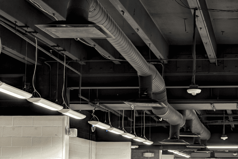 Exposed black ventilation ducts and hanging fluorescent lights on an industrial-style ceiling above a white brick wall.