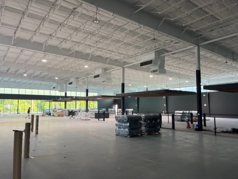Interior of a large, unfinished warehouse or commercial space with high ceilings, stacked materials, and construction equipment.