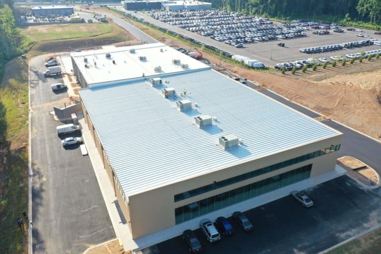Aerial view of a large industrial warehouse with a metal roof, adjacent parking lot, and several parked cars, surrounded by greenery and other buildings.