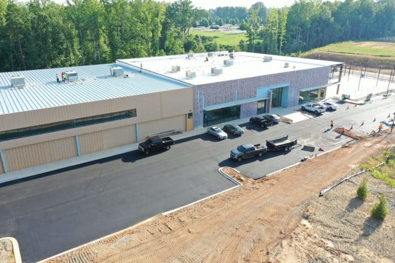 Aerial view of a modern commercial building under construction with newly paved parking lot, several vehicles parked, and workers present on site.