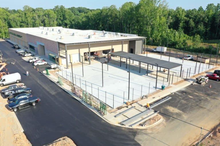 Aerial view of a construction site featuring a large industrial building with a partially completed exterior and adjacent parking area, surrounded by trees.