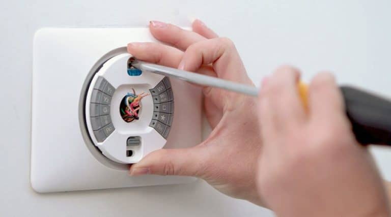 A person uses a screwdriver to install or repair a smart thermostat on a white wall, exposing internal wires and components.