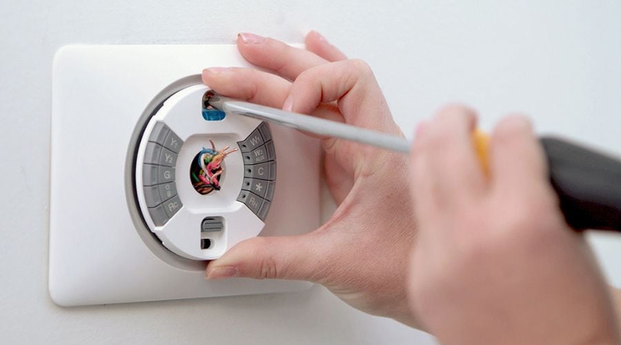 A person uses a screwdriver to install or repair a smart thermostat on a white wall, exposing internal wires and components.