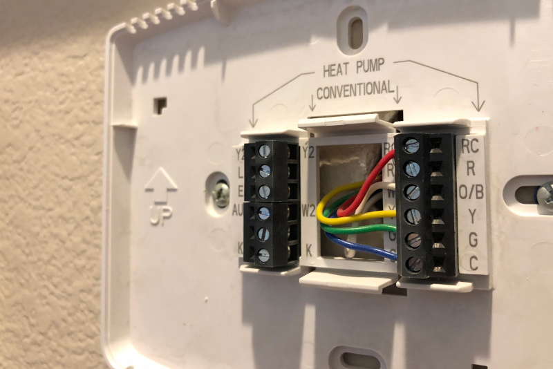 A close-up of a wall-mounted thermostat base showing exposed wiring and labeled connection terminals for a heat pump and conventional system.