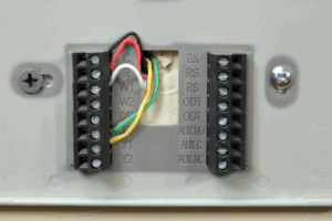 Close-up of a thermostat wiring panel showing exposed colored wires connected to labeled terminals, with screws securing the panel to the wall.