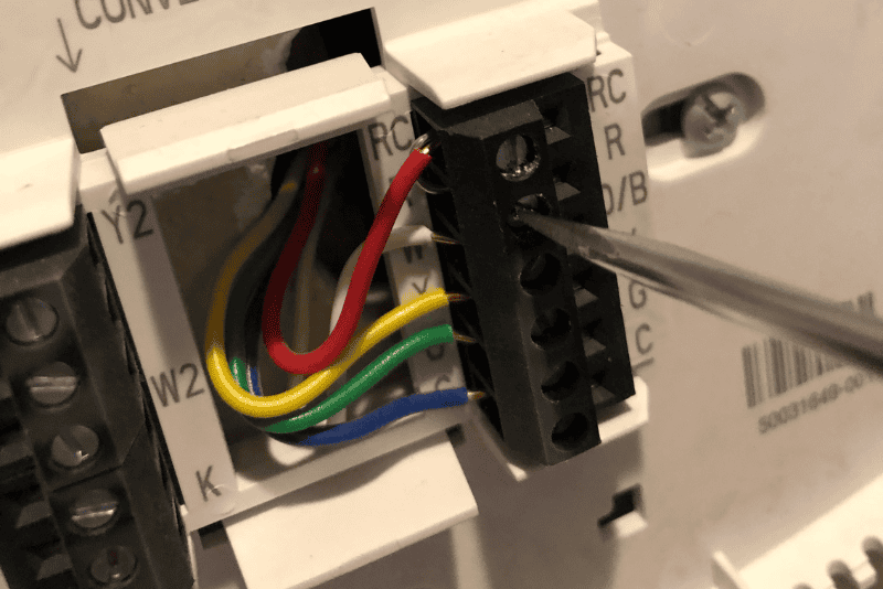 Close-up of a screwdriver adjusting wiring connections on a thermostat terminal block with colored wires attached.